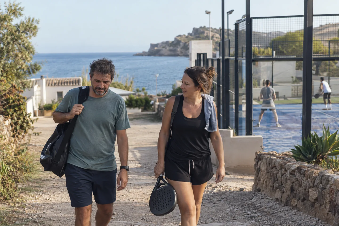 Two adults walk beside a coastal club with a sports bag and padel racket.